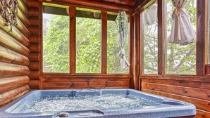 Interior de una cabaña de madera con jacuzzi, ventanas amplias y vista a la vegetación.