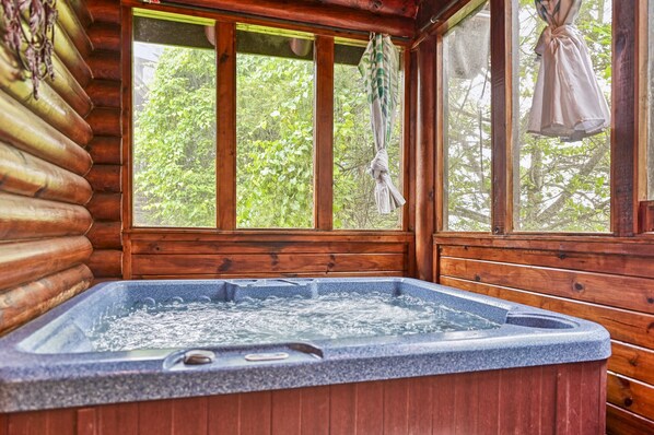 Interior de una cabaña de madera con jacuzzi, ventanas amplias y vista a la vegetación.