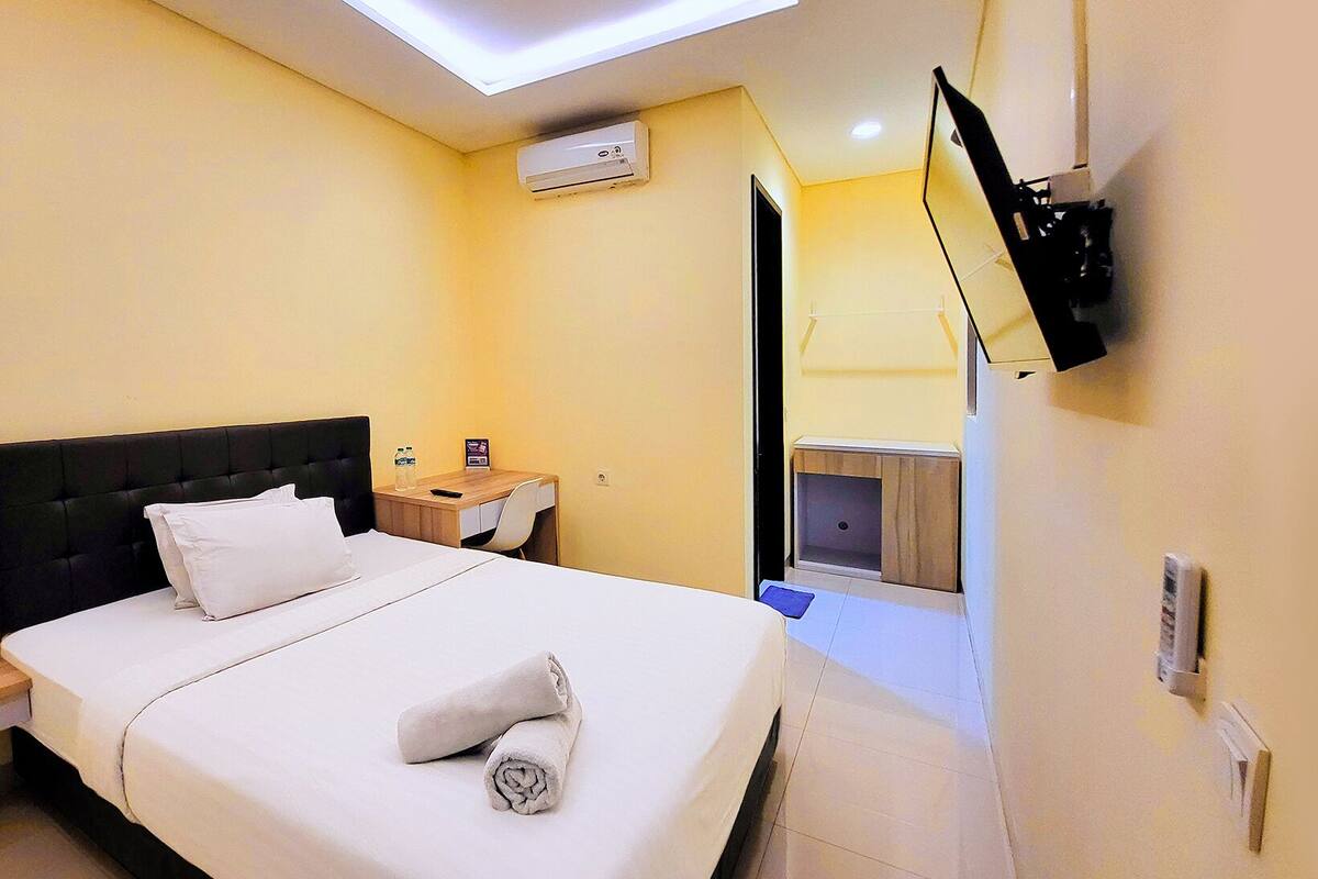 A hotel room with a bed, a bedside table with a bottle and remote control, a wall-mounted TV, and a small wooden cabinet.