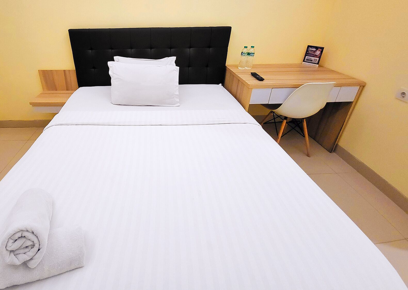 A hotel room with a bed, a desk, a chair, and a small table with a bottle of water and a remote control.
