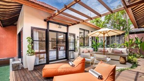 A sunlit patio area with wooden flooring, two orange cushioned chairs, a white sofa with cushions, and a large umbrella.