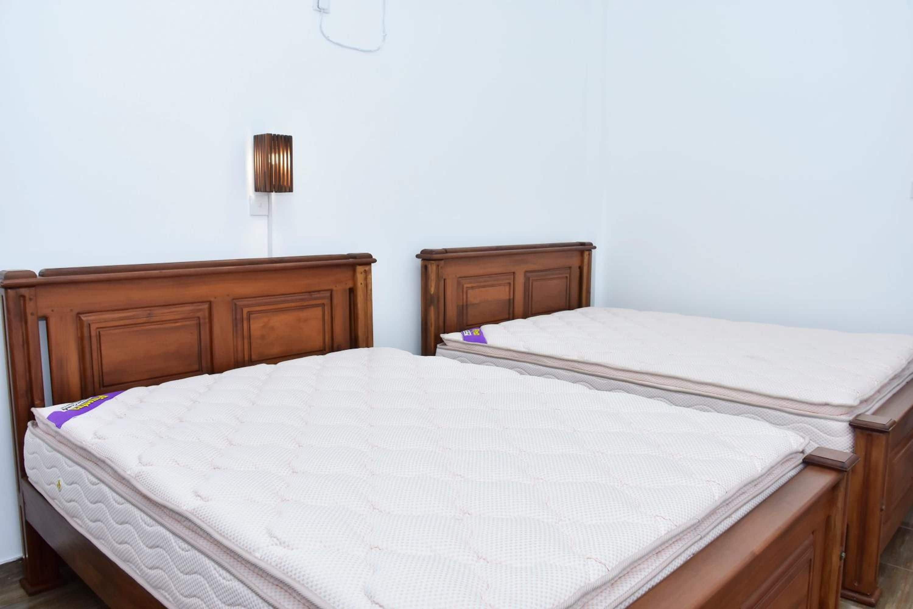 Two wooden beds with white mattresses in a room with a white wall and a wall-mounted light fixture.