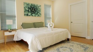 A neatly made bed with a white comforter, a green pillow, and a decorative swan-shaped pillow. There is a nightstand with a lamp and a small plant beside the bed. A large green floral artwork hangs on the wall above the bed. - 🔥WALK 2 BEACH❤️ | SLEEPS 6 W/ W+D (Biloxi)