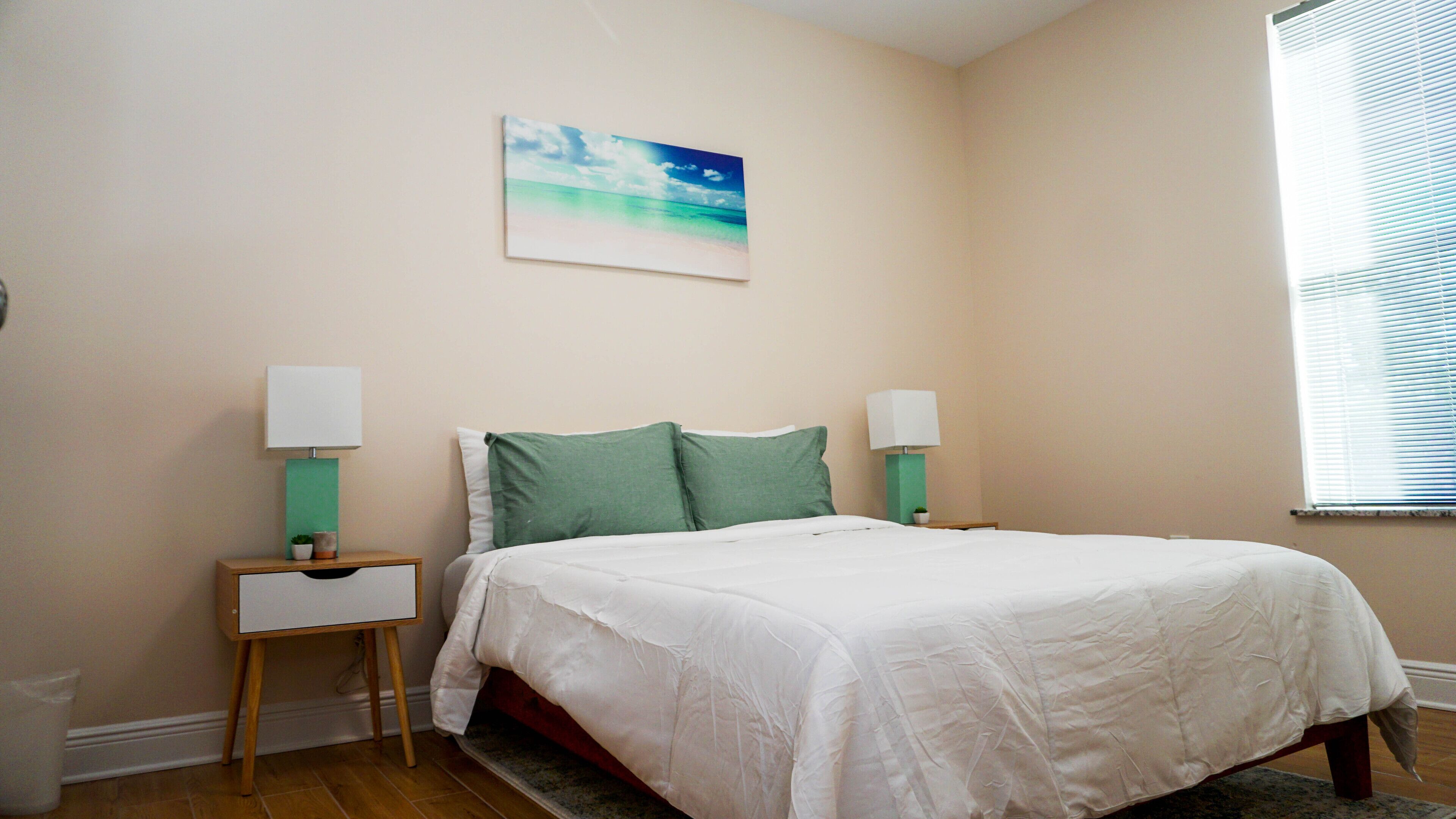 Une chambre à coucher comprenant un lit, deux tables de chevet, une commode, un tableau accroché au mur et une fenêtre avec des stores.