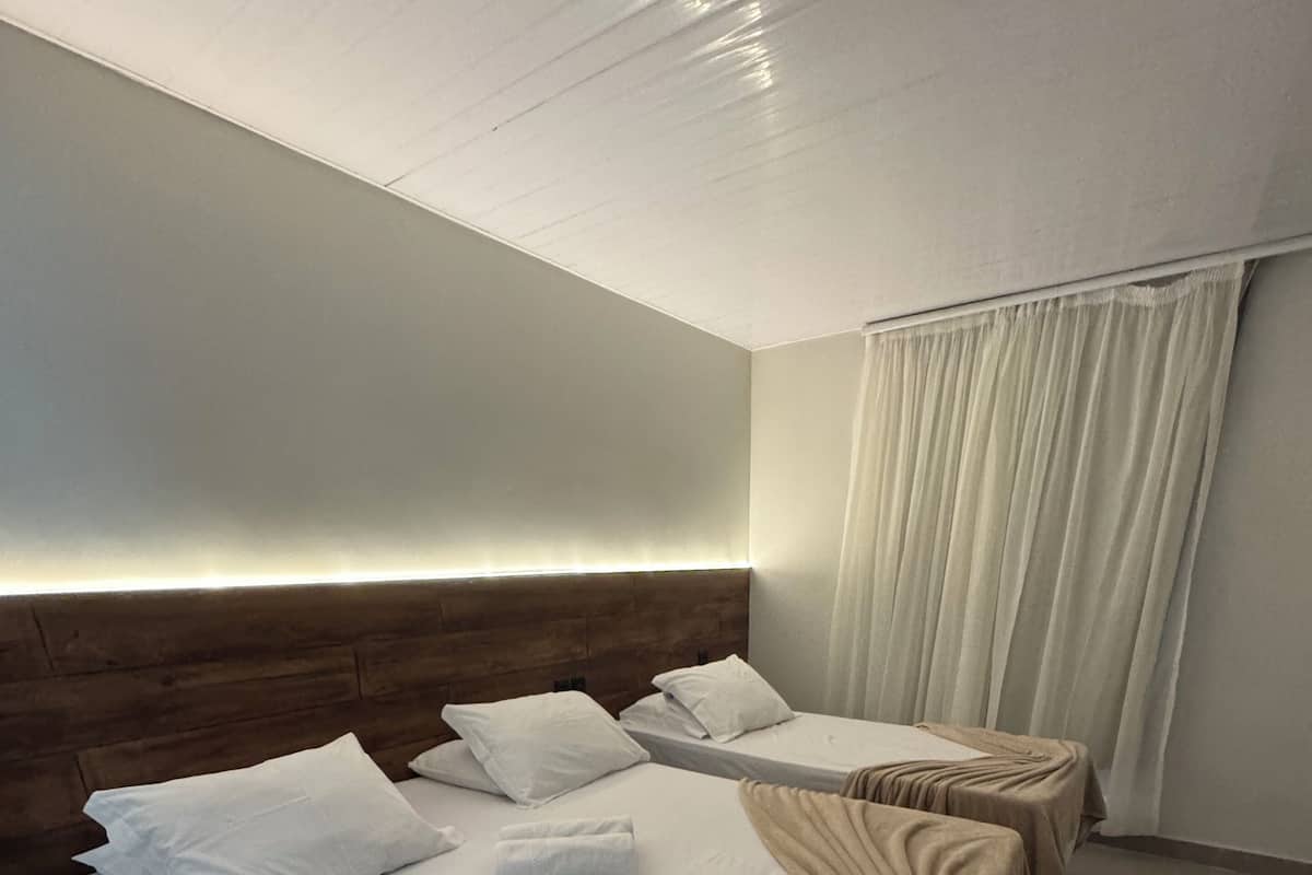 Ein Hotelzimmer mit zwei Betten, jeweils mit weißer Bettwäsche und einer braunen Tagesdecke. Das Zimmer verfügt über ein Holz-Kopfteil, ein großes Fenster mit weißen Vorhängen und einen Flachbildfernseher auf der rechten Seite.