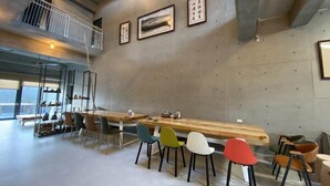 A modern dining area with a long wooden table and colorful chairs, a concrete wall with framed pictures, and a mezzanine level with a railing. - Green House Homestay (Magong)
