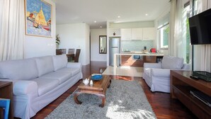 A modern living room with a sofa, coffee table, and TV. A kitchen is visible in the background. - Kata gardens 2 bedroom near Kata beach 3B (Karon)