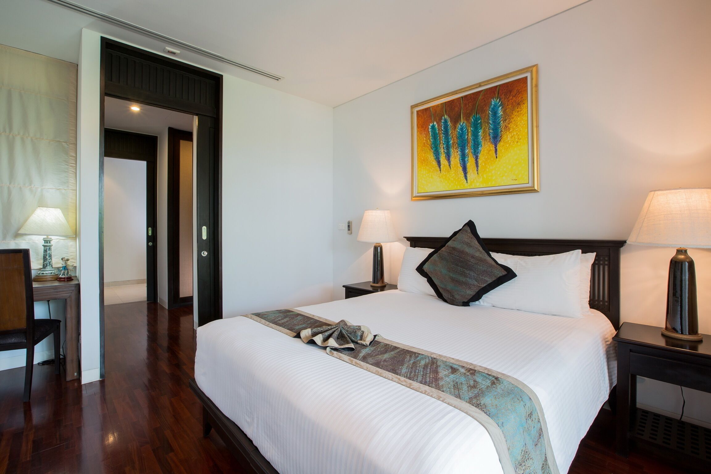 A hotel room with a large bed, two bedside tables with lamps, a painting on the wall, and a door leading to another room.