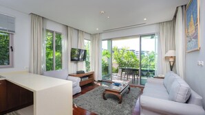A modern living room with a sofa, a coffee table, and a TV. There is a balcony with a view of greenery. - Kata gardens 2 bedroom near Kata beach 3B (Karon)