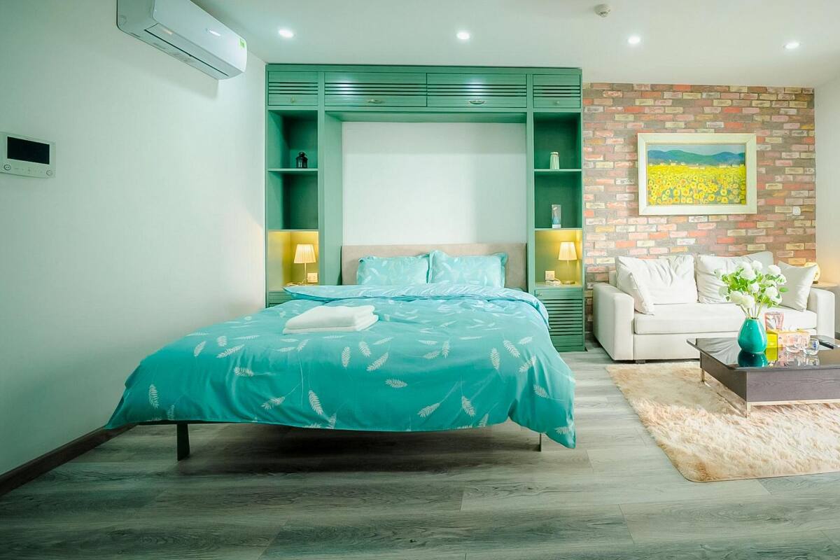 A modern bedroom with a bed, a sofa, a coffee table, and a large window with a view of buildings and greenery.