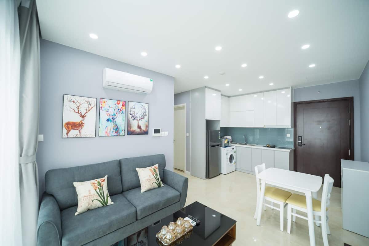 A modern living room with a grey sofa, a dining table with chairs, and a kitchen area with white cabinets and appliances.