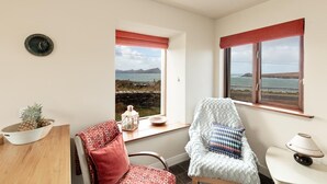 A cozy room with a view of the sea, a wooden table with a pineapple, a chair with patterned cushions, and a lamp.