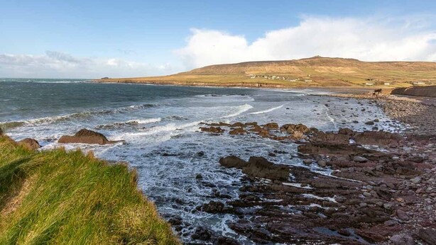 A coastal landscape with rocky shores, grassy areas, and distant hills under a clear sky.