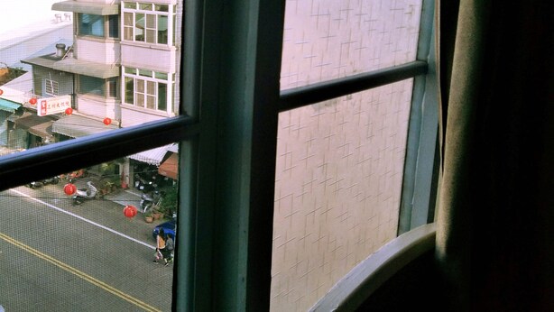 A view through a window with a grid pattern on the wall, a building with multiple floors, and a street scene with parked vehicles and people. - Pon Kan Inn (Beigang)