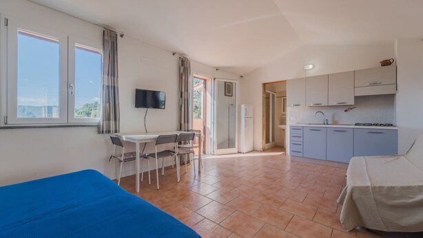 A room with a bed, a desk with a chair, a TV, a kitchen area, and a sliding door leading to an outdoor space. - APP.TI NORSI - NORSI - MONO (Capoliveri)