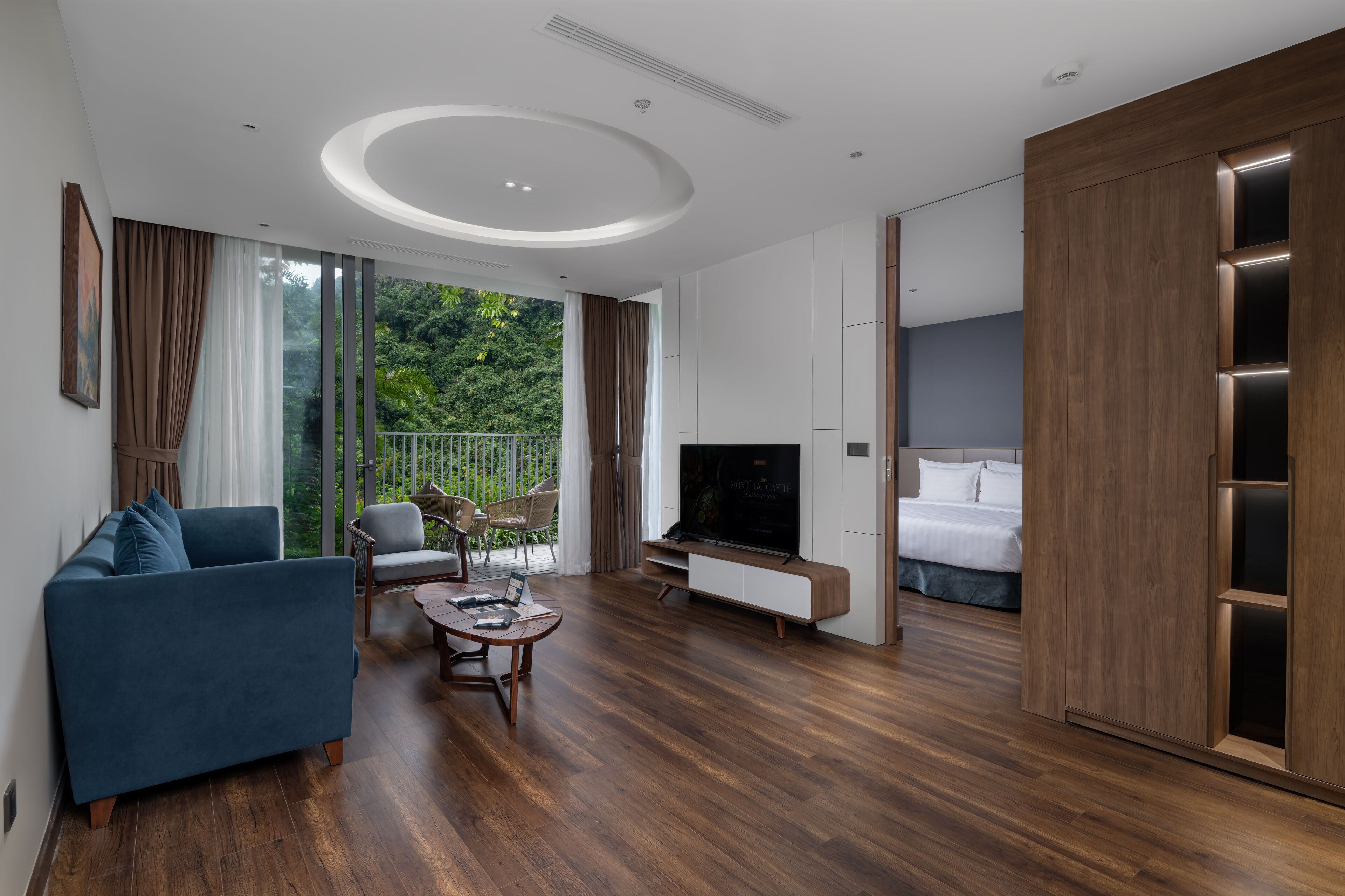 A modern living room with a sofa, TV, and a balcony with a view of greenery.