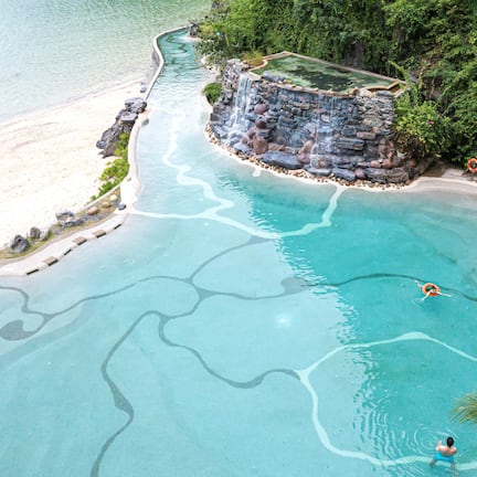 An aerial view of a resort with a large swimming pool, a beach, and lush greenery.