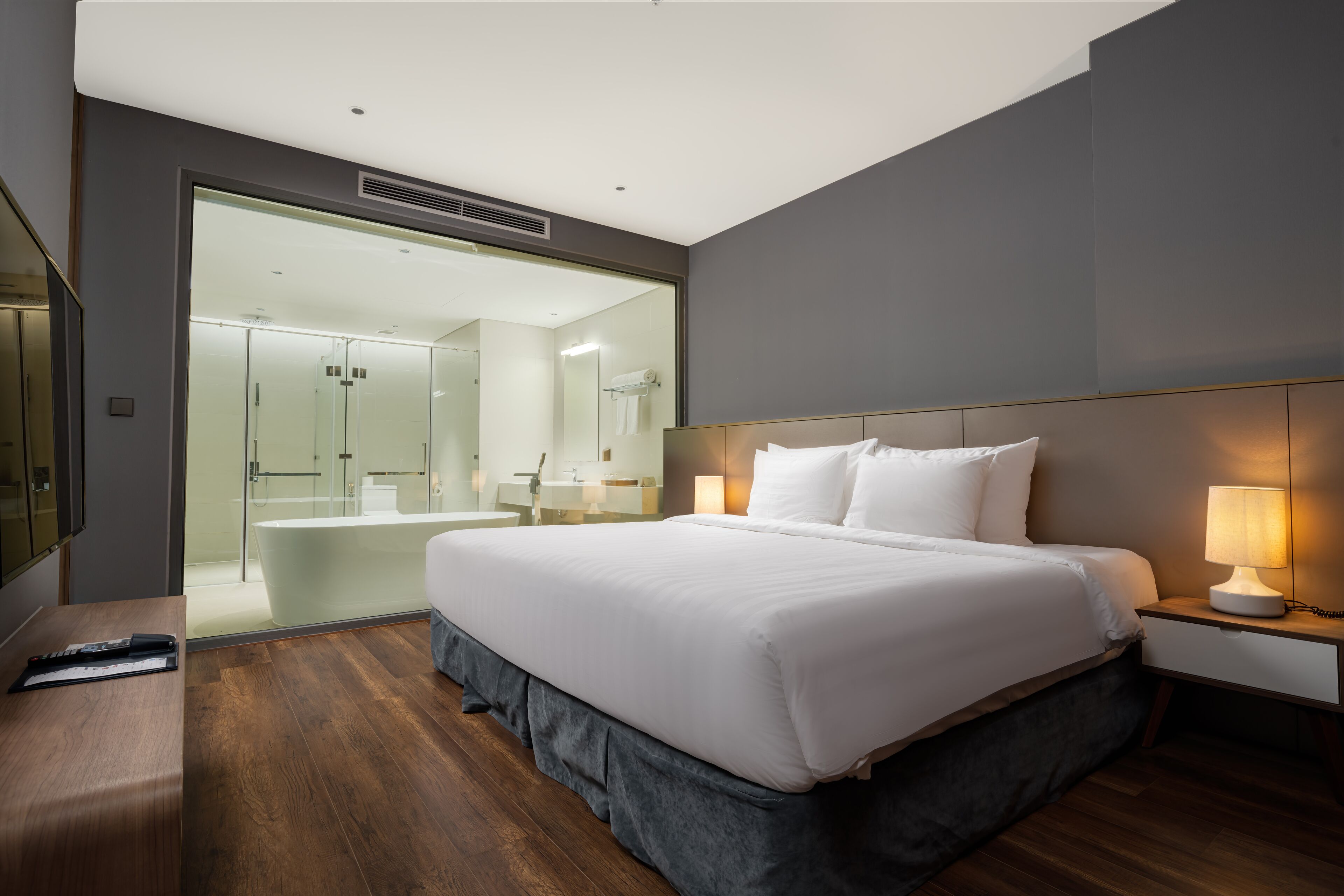 Ein modernes Hotelzimmer mit einem großen Bett, einem Nachttisch und einem durch eine Glastür einsehbaren Badezimmer.