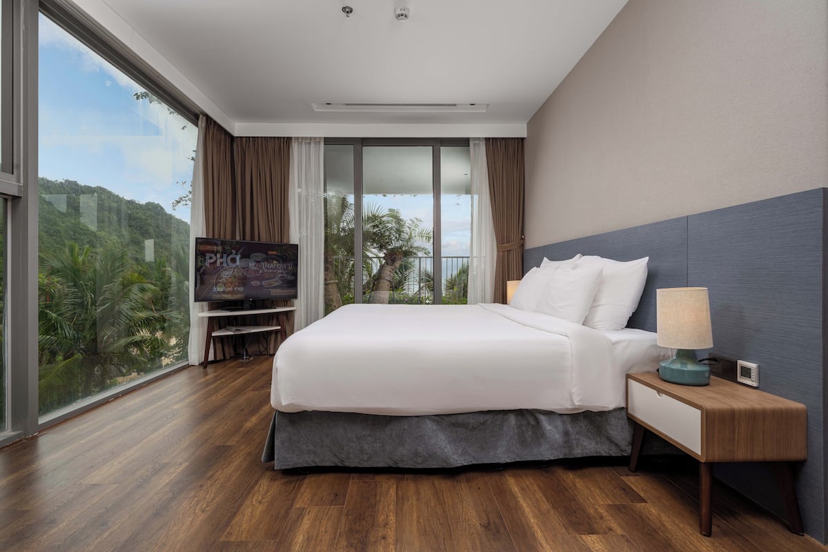 Une chambre d’hôtel moderne dotée d’un grand lit, d’une télévision, d’une table de chevet avec une lampe et offrant une vue sur un paysage luxuriant grâce à de grandes fenêtres.