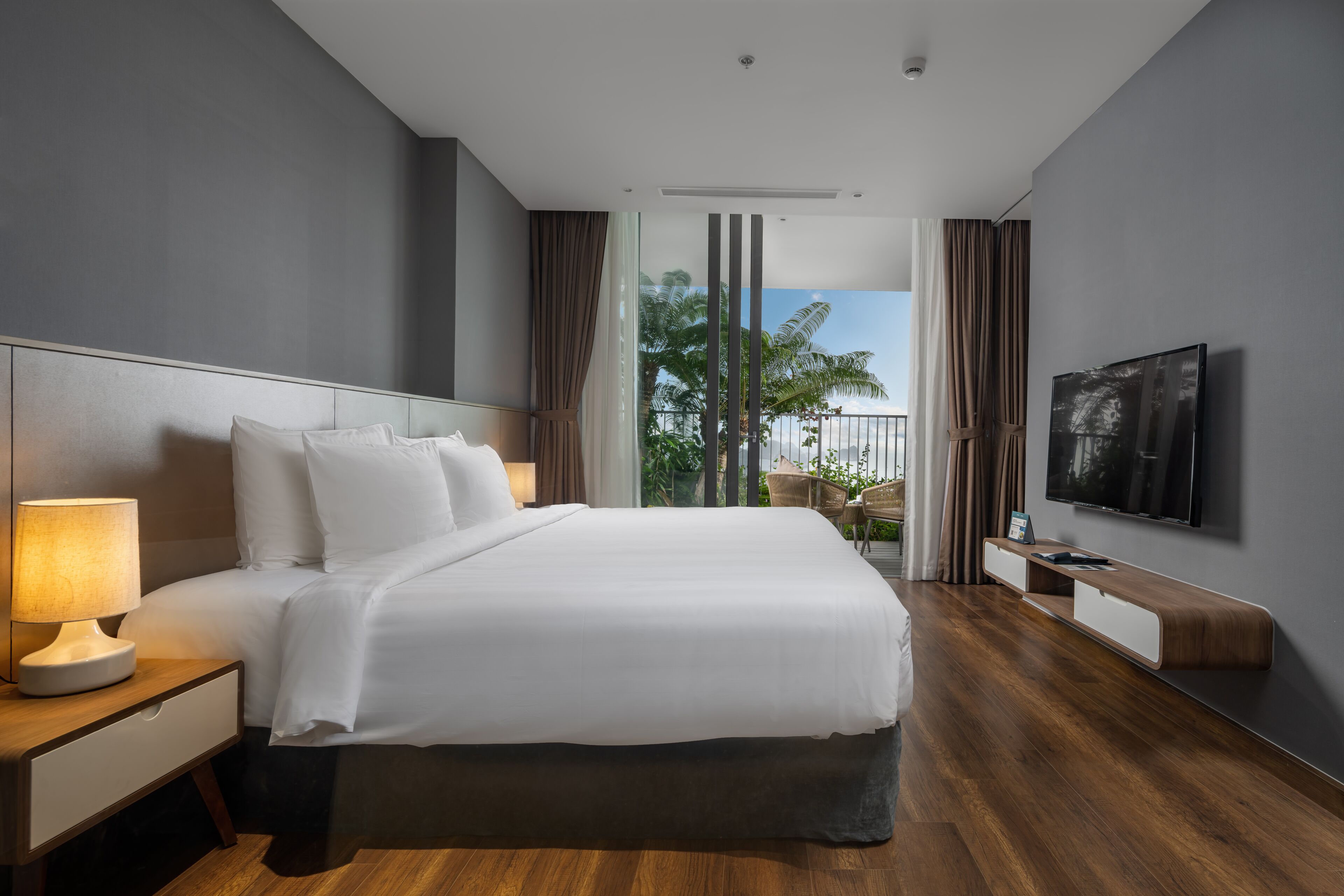 Ein modernes Hotelzimmer mit einem großen Bett, einem Nachttisch mit Lampe, einem an der Wand montierten Fernseher und einem Balkon mit Blick auf Palmen.
