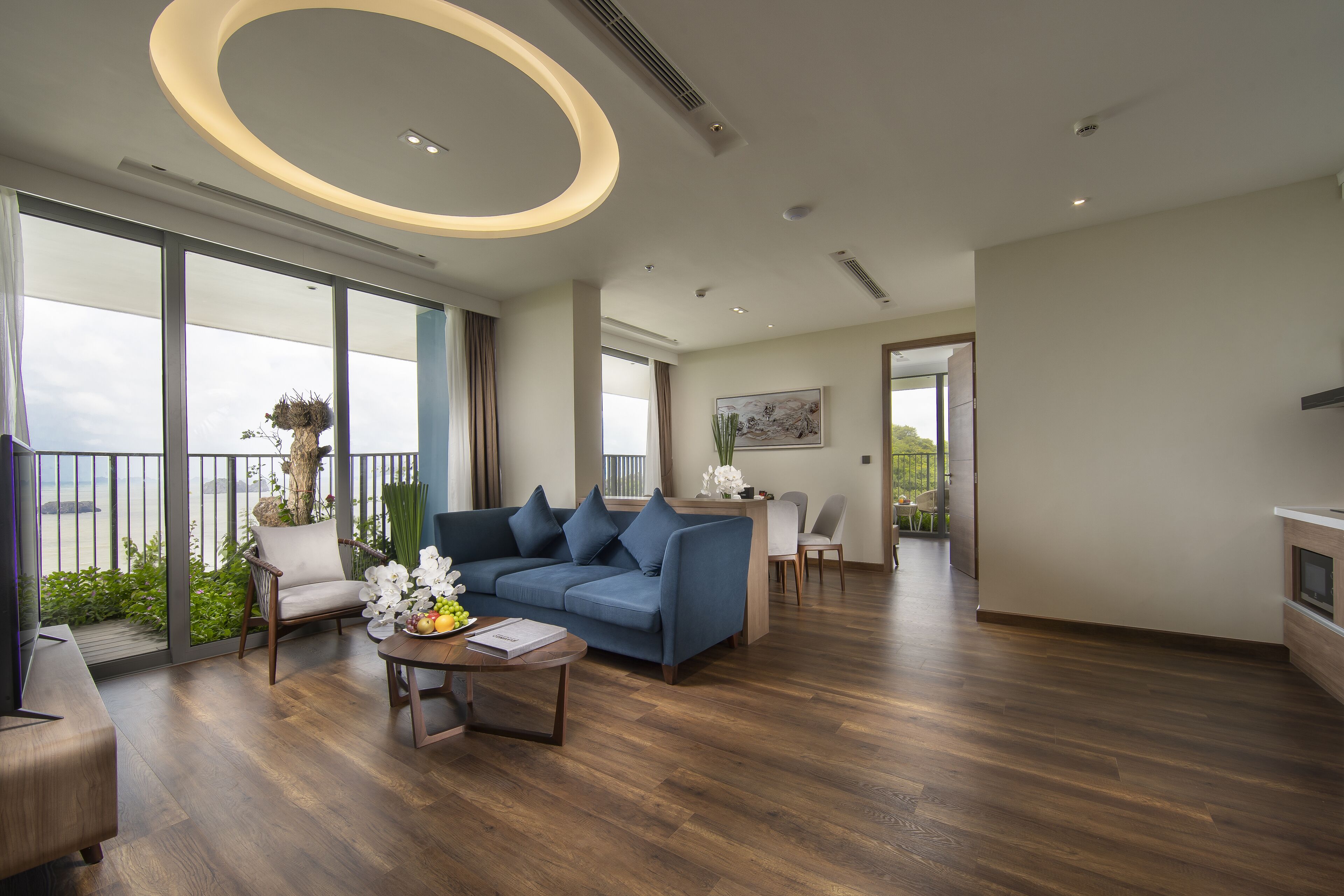 Uma sala de estar ampla com sofá azul, piso de madeira e grandes janelas com vista para o verde e uma varanda.
