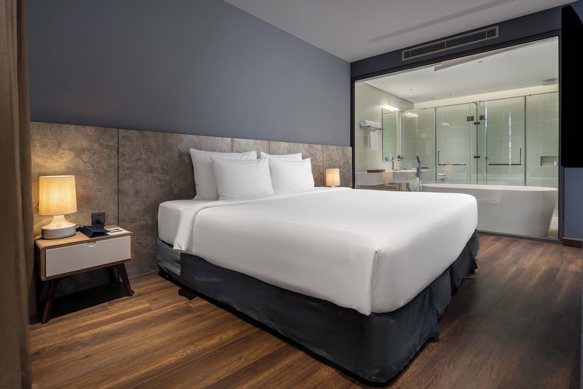 Une chambre d’hôtel moderne avec un grand lit, une table de chevet avec une lampe et une salle de bain fermée par une paroi de verre.