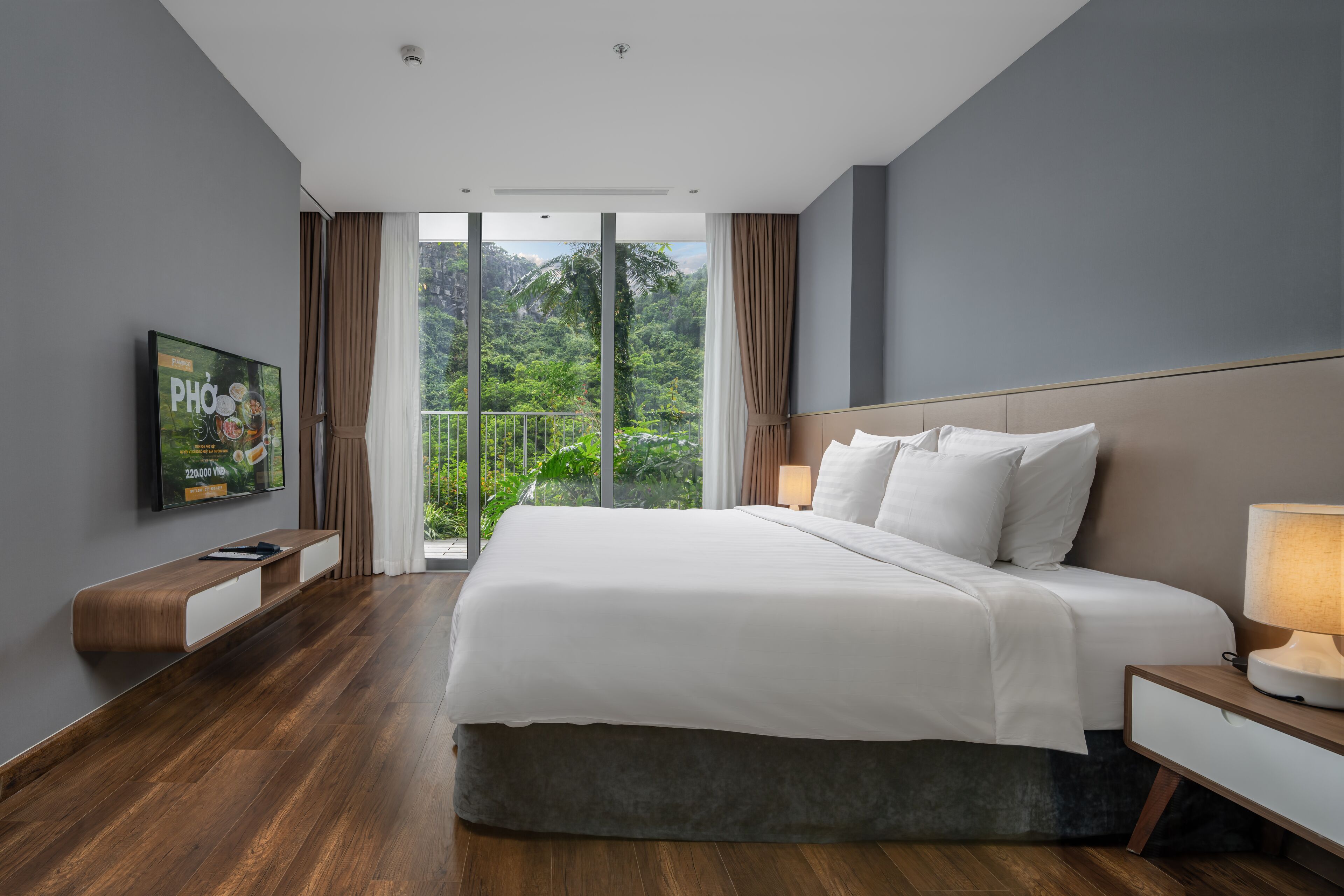 Een moderne hotelkamer met een groot bed, een flatscreen televisie, een houten console tafel en een balkon met uitzicht op groen.