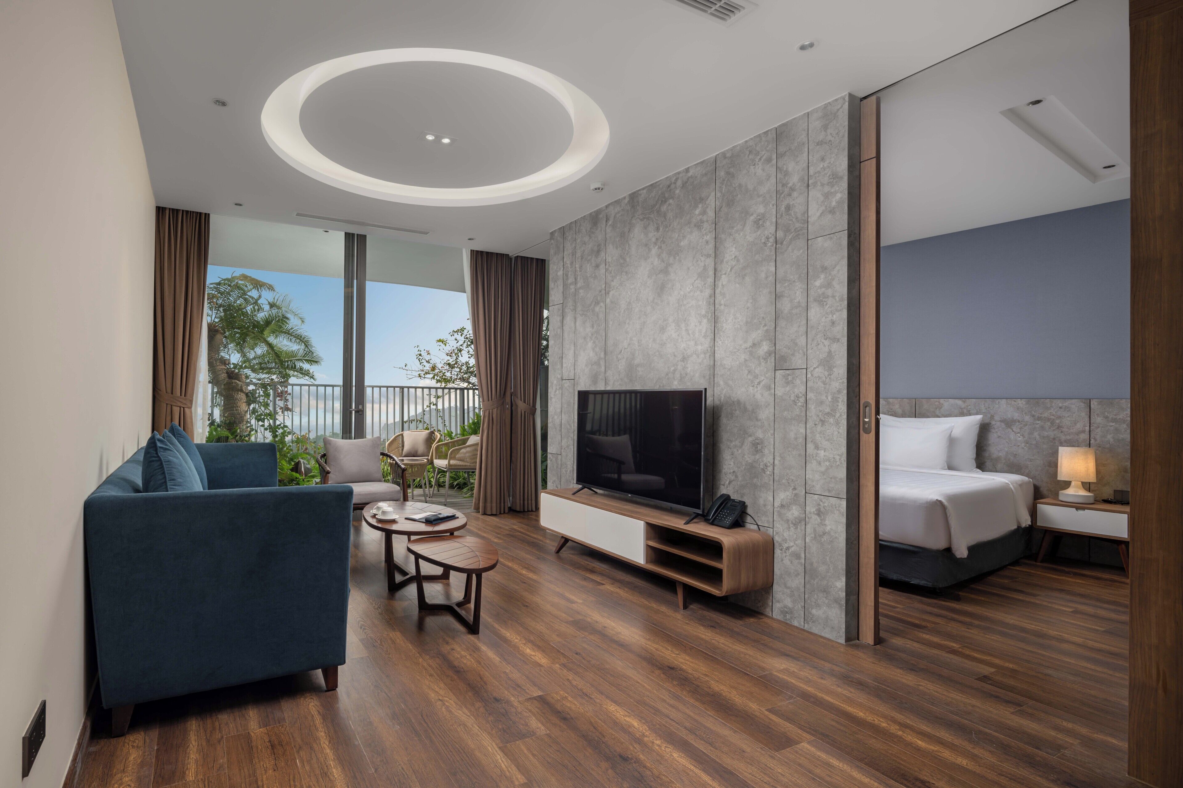 A modern hotel room with a large TV, a sofa, a small coffee table, and a bed with a nightstand.