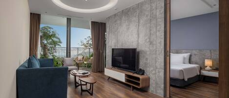 A modern hotel room with a large TV, a sofa, a small coffee table, and a bed with a nightstand.