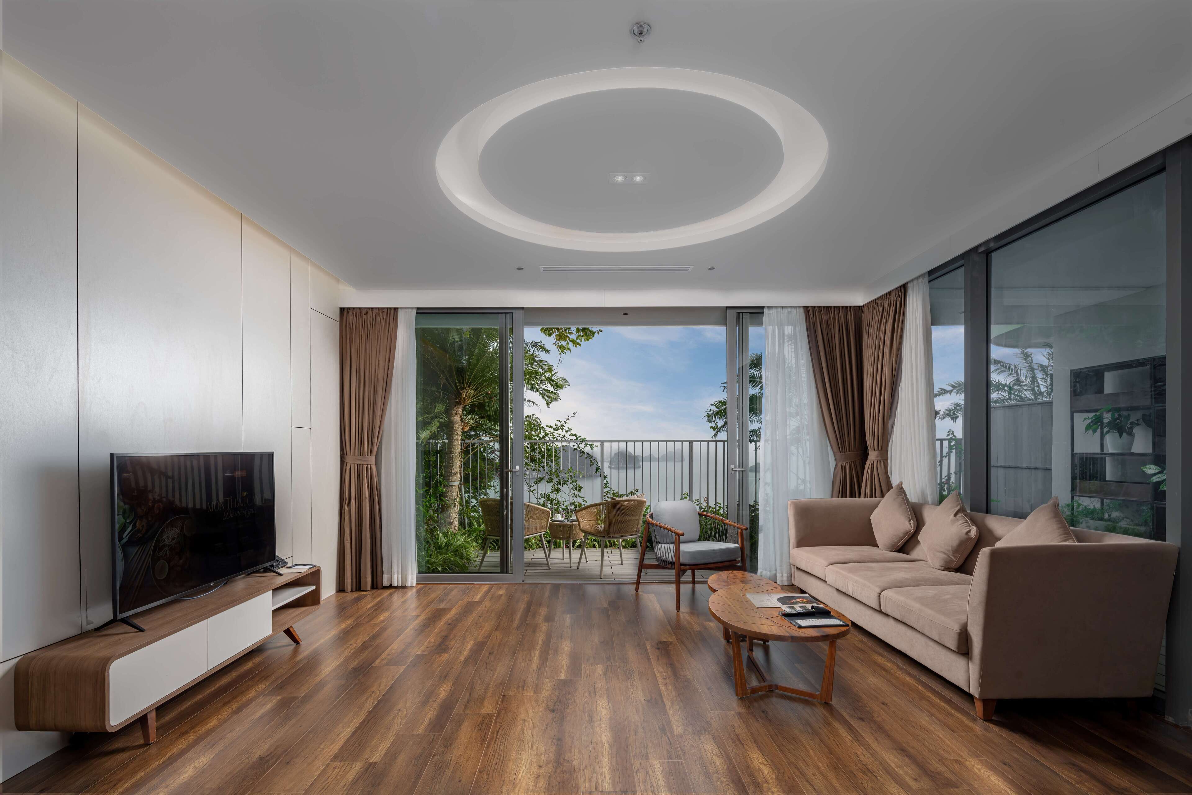 A modern living room with a flat-screen TV, wooden flooring, a sofa, a coffee table, and a sliding glass door leading to an outdoor area with plants and a balcony.