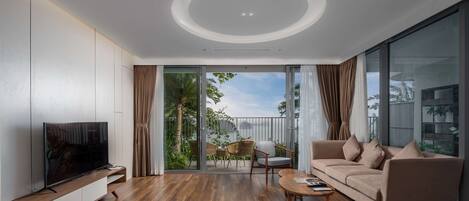 A modern living room with a flat-screen TV, wooden flooring, a sofa, a coffee table, and a sliding glass door leading to an outdoor area with plants and a balcony.