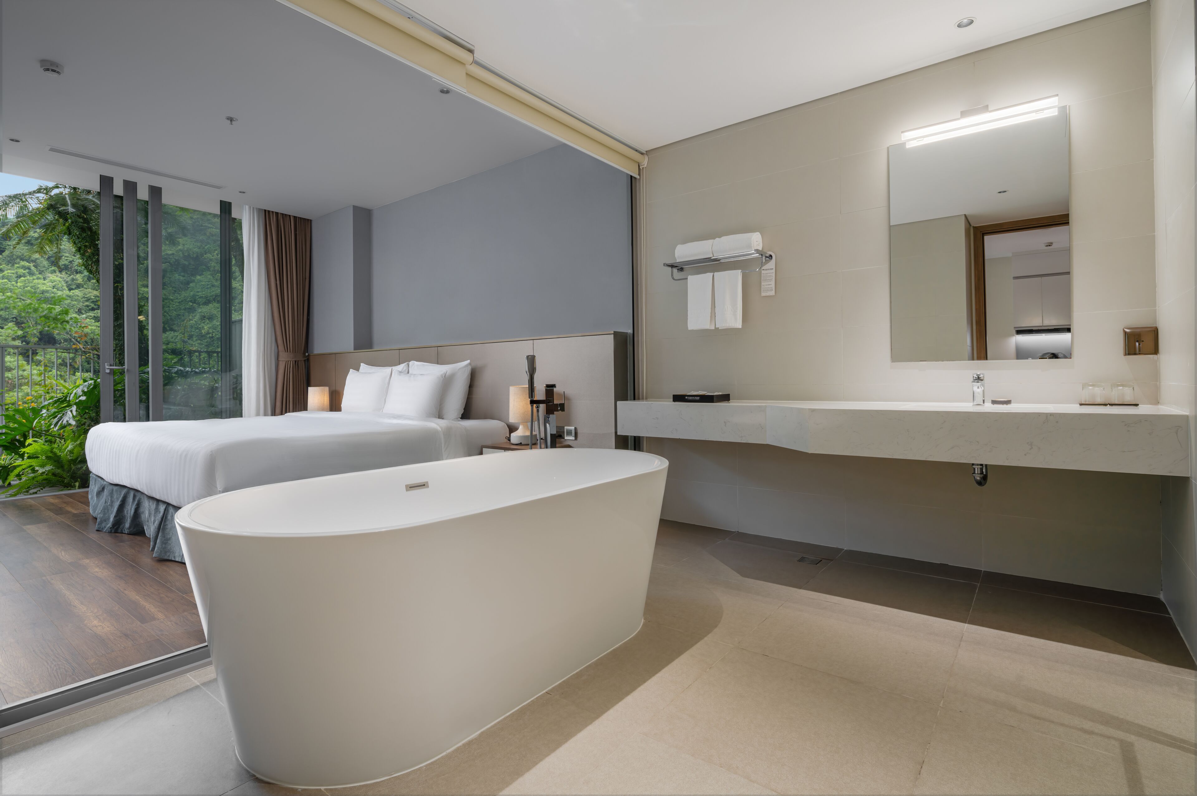 A modern hotel room with a large bathtub, a double bed, a vanity with a mirror, and a view of greenery through large windows.