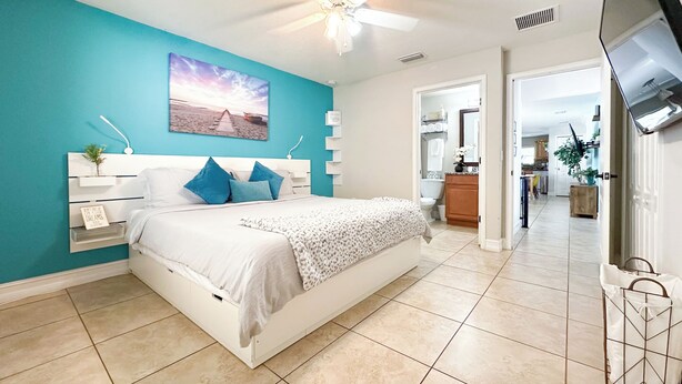 Une chambre à coucher avec un lit, un ventilateur de plafond, un téléviseur fixé au mur et une salle de bain visible à travers une porte ouverte.