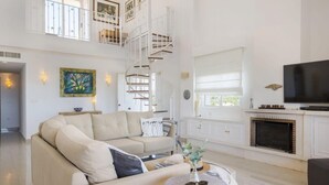 A spacious living room with a beige sofa, a round coffee table with decorative items, a fireplace, and a TV.