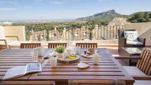 A wooden table set with a book, glasses, and a small plant on a balcony overlooking a city and mountains.