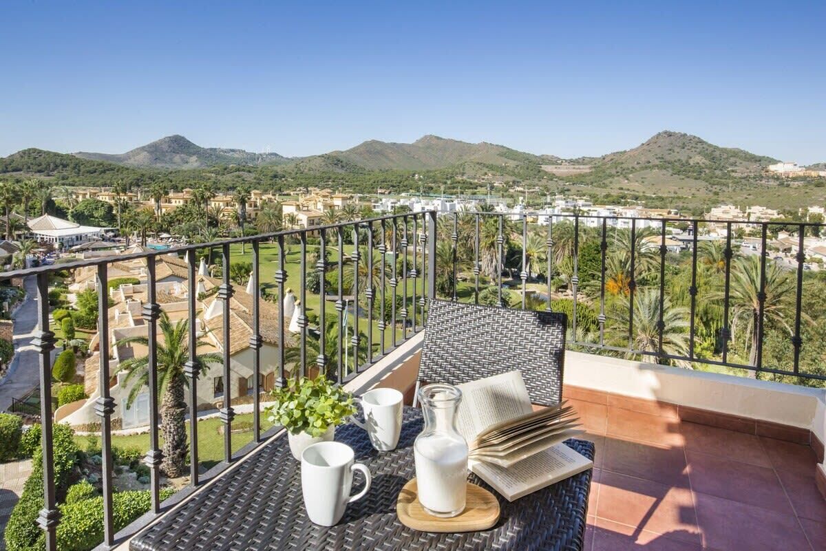 A balcony with a view of a residential area, a table with a book, a glass pitcher, and two white mugs.