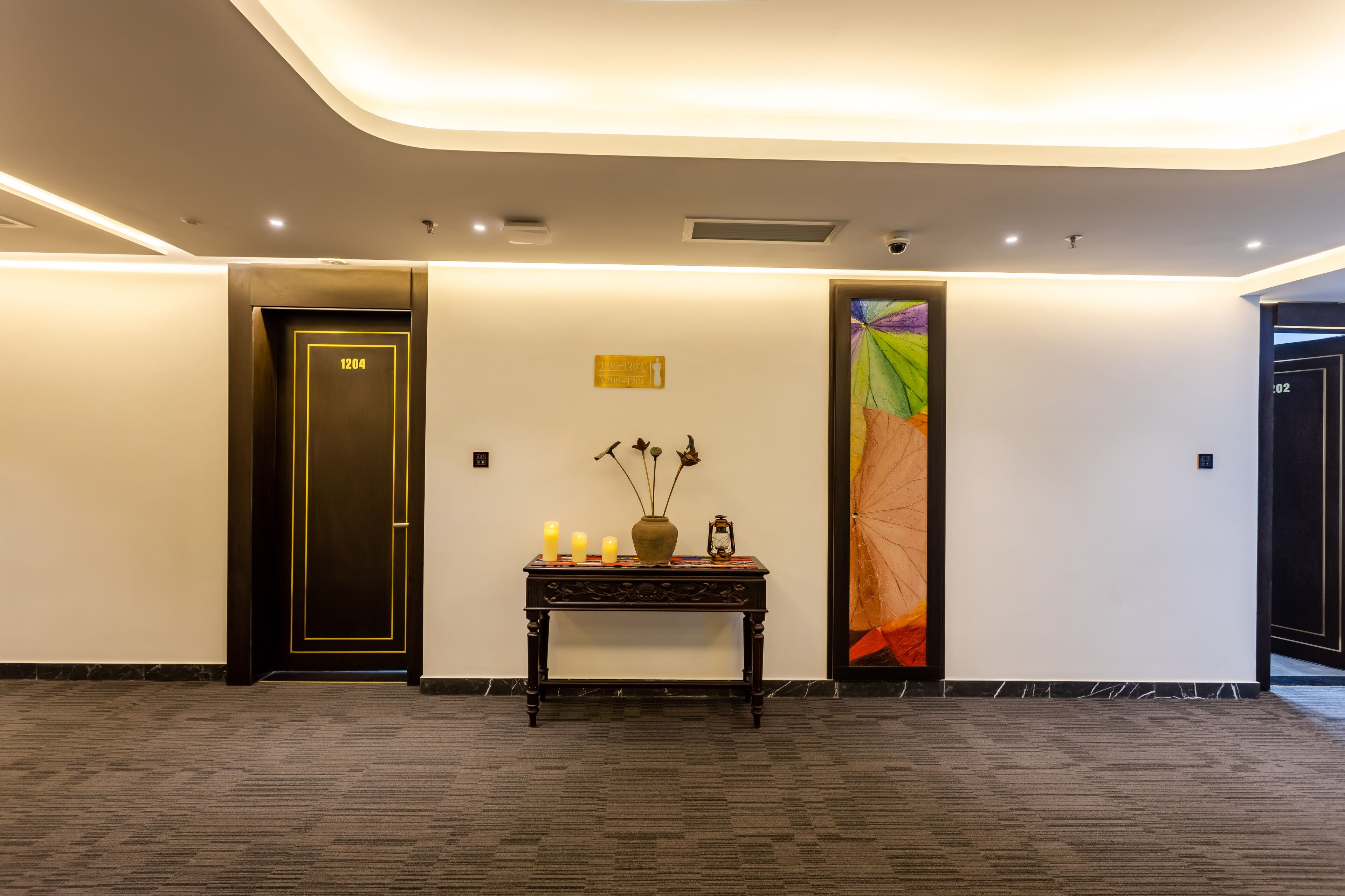 A modern hotel lobby with a dark wooden door marked "1204", a console table with candles and a vase, and a colorful abstract painting on the wall.