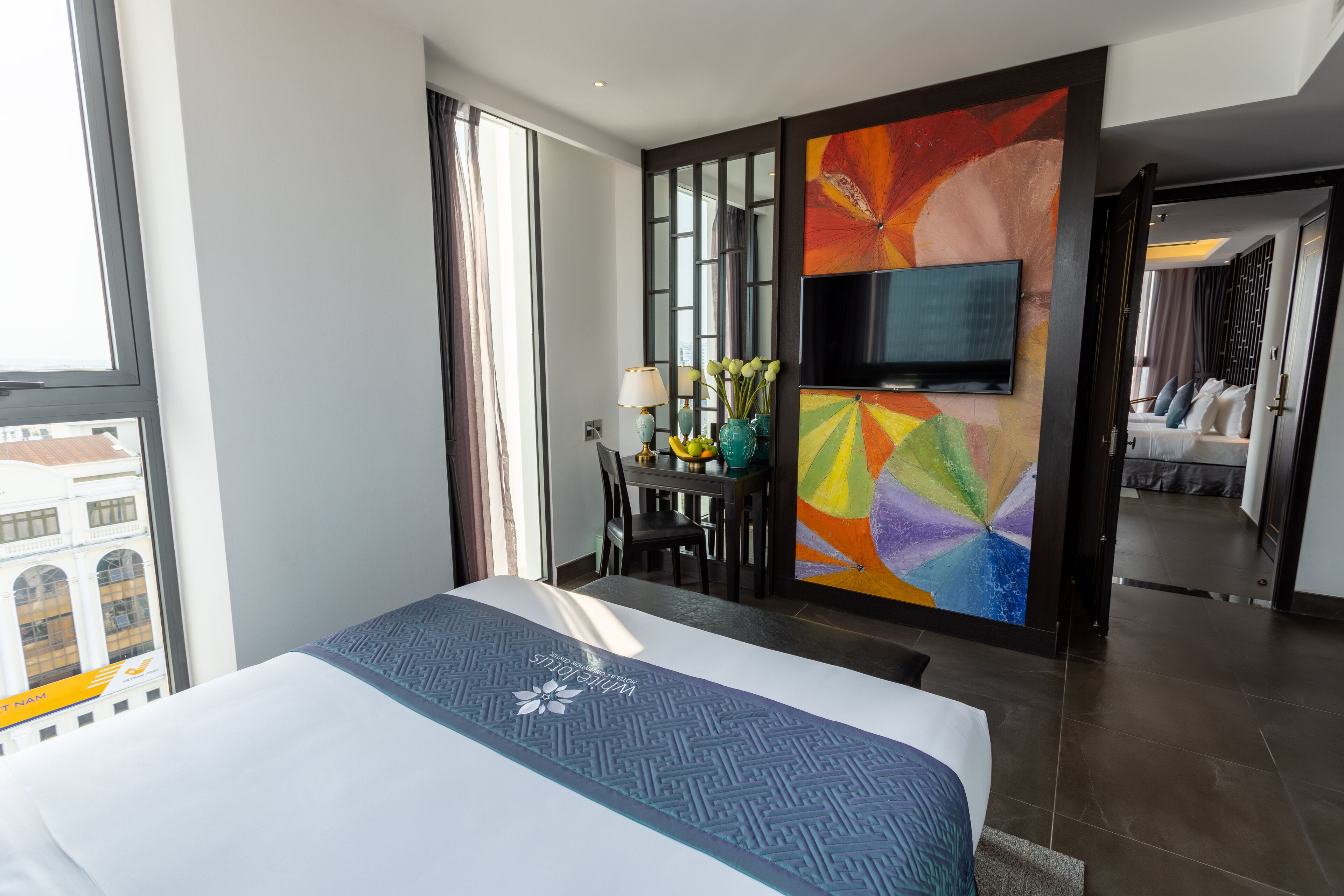 A hotel room with a bed, a TV, a dining area with a table and chairs, and a colorful abstract painting on the wall.