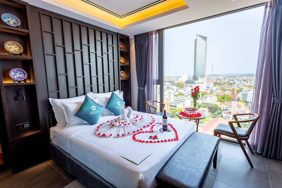 A hotel room with a bed, a bedside table with a wine bottle, a chair, and a view of a cityscape through large windows.