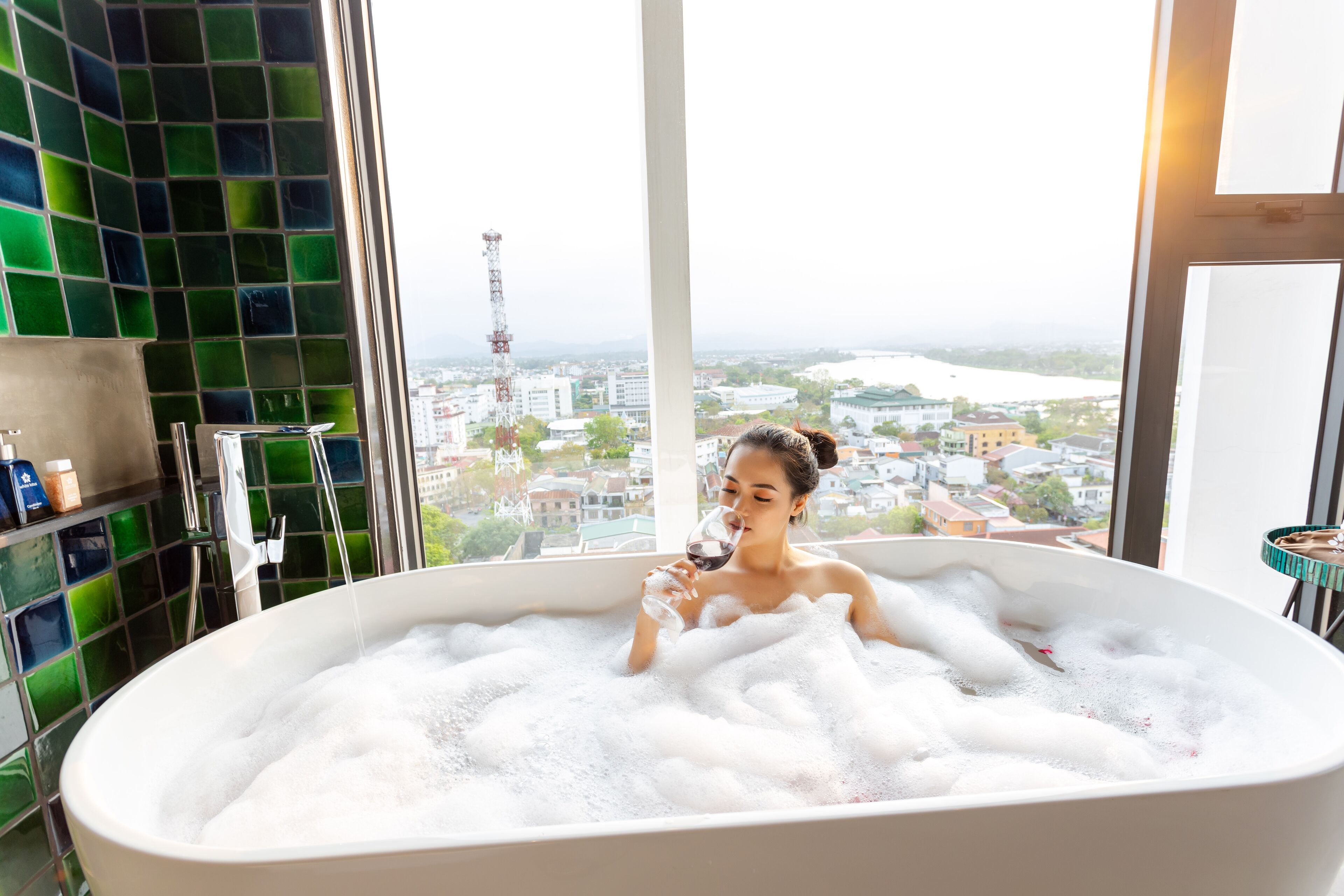A woman in a bathtub with a city view, a green-tiled wall, and a window.