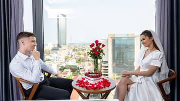 A couple seated at a table with a cake and roses, overlooking a cityscape through large windows.