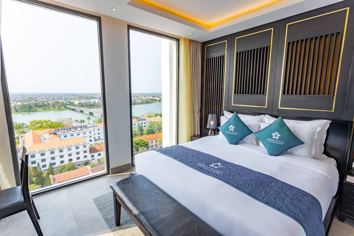 A hotel room with a large bed, two pillows, a bench, and a view of a river and buildings.