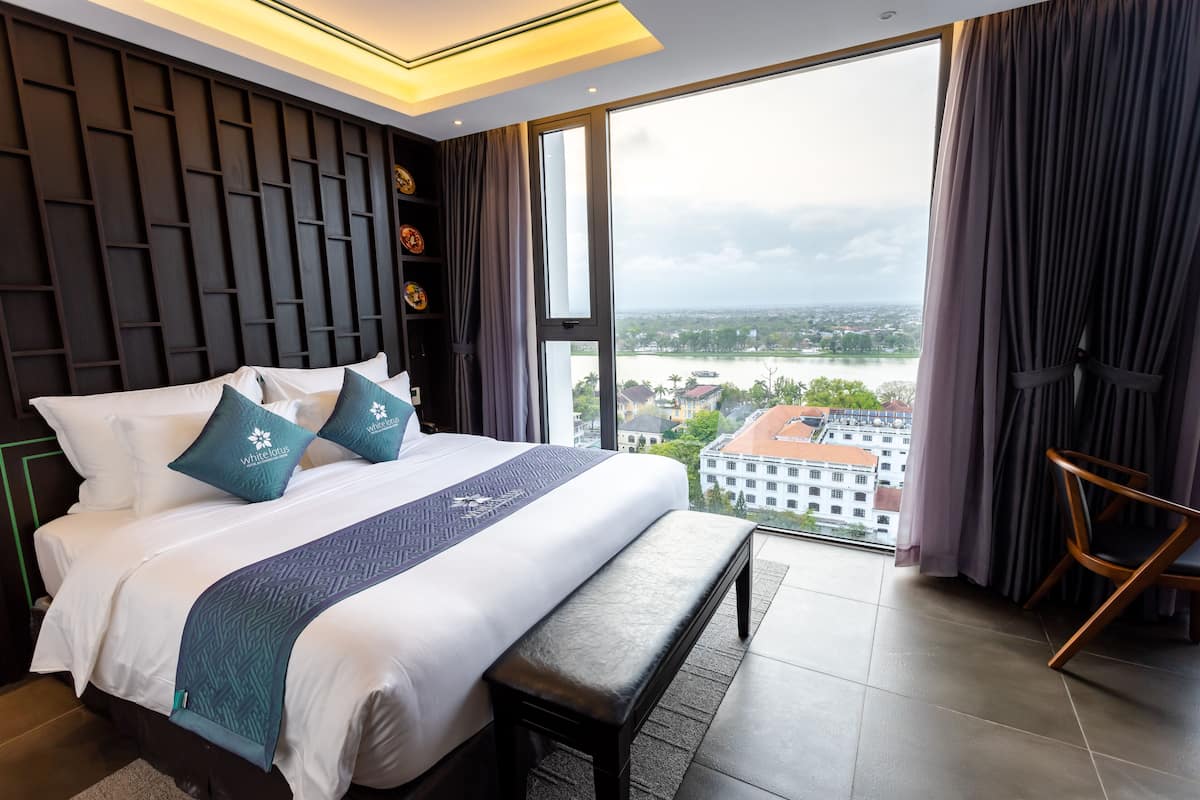 A modern hotel room with a large bed, a bench, a view of the city, and a wooden chair.