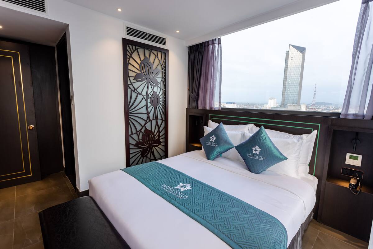 A hotel room with a large bed, a view of a cityscape, a decorative wall panel, and a modern control panel.
