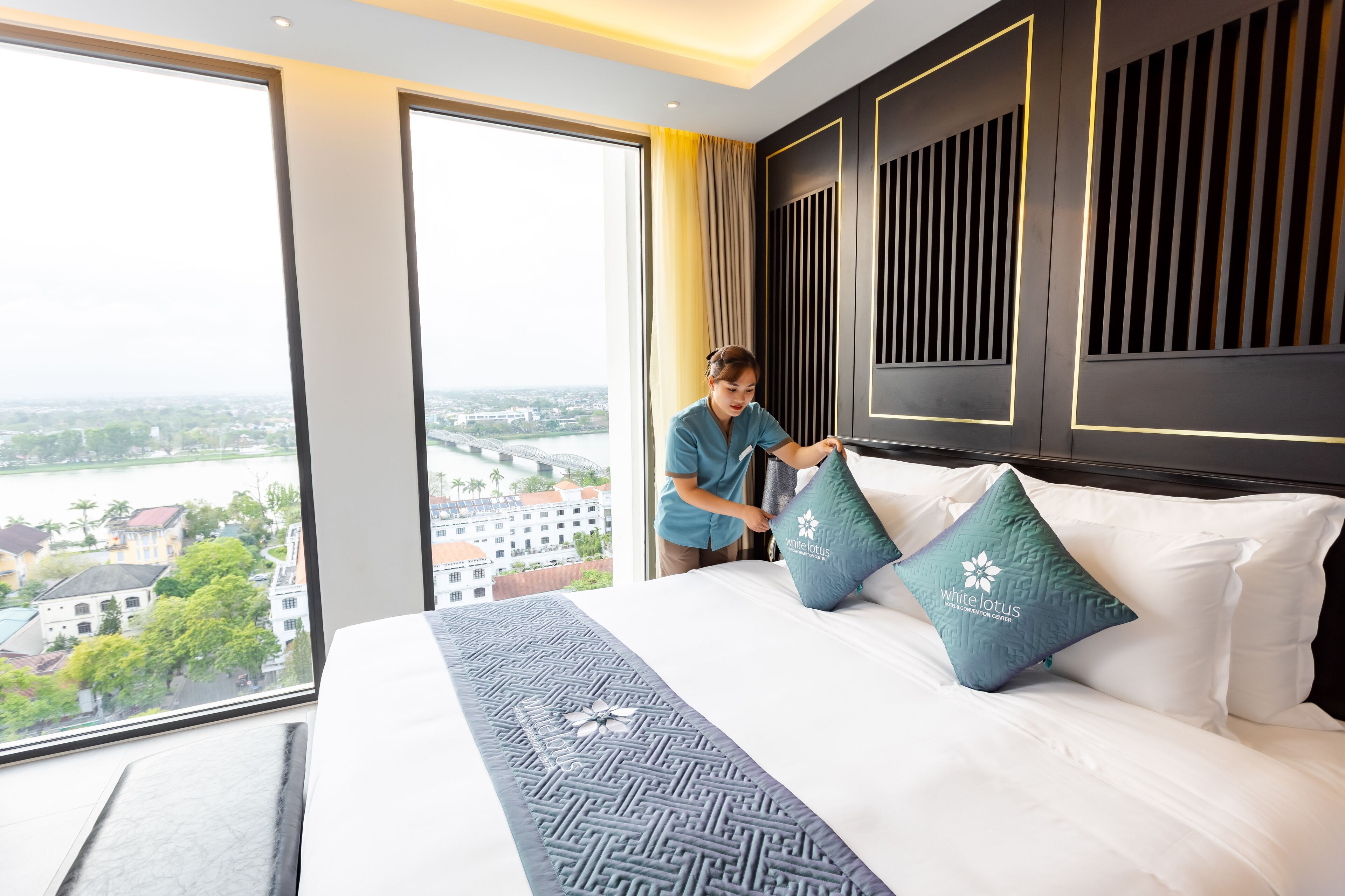 A hotel room with a large bed, a view of a cityscape through the window, and a person adjusting a pillow.