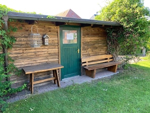 A rustic wooden building with a green door, two benches, and a birdhouse on the wall.