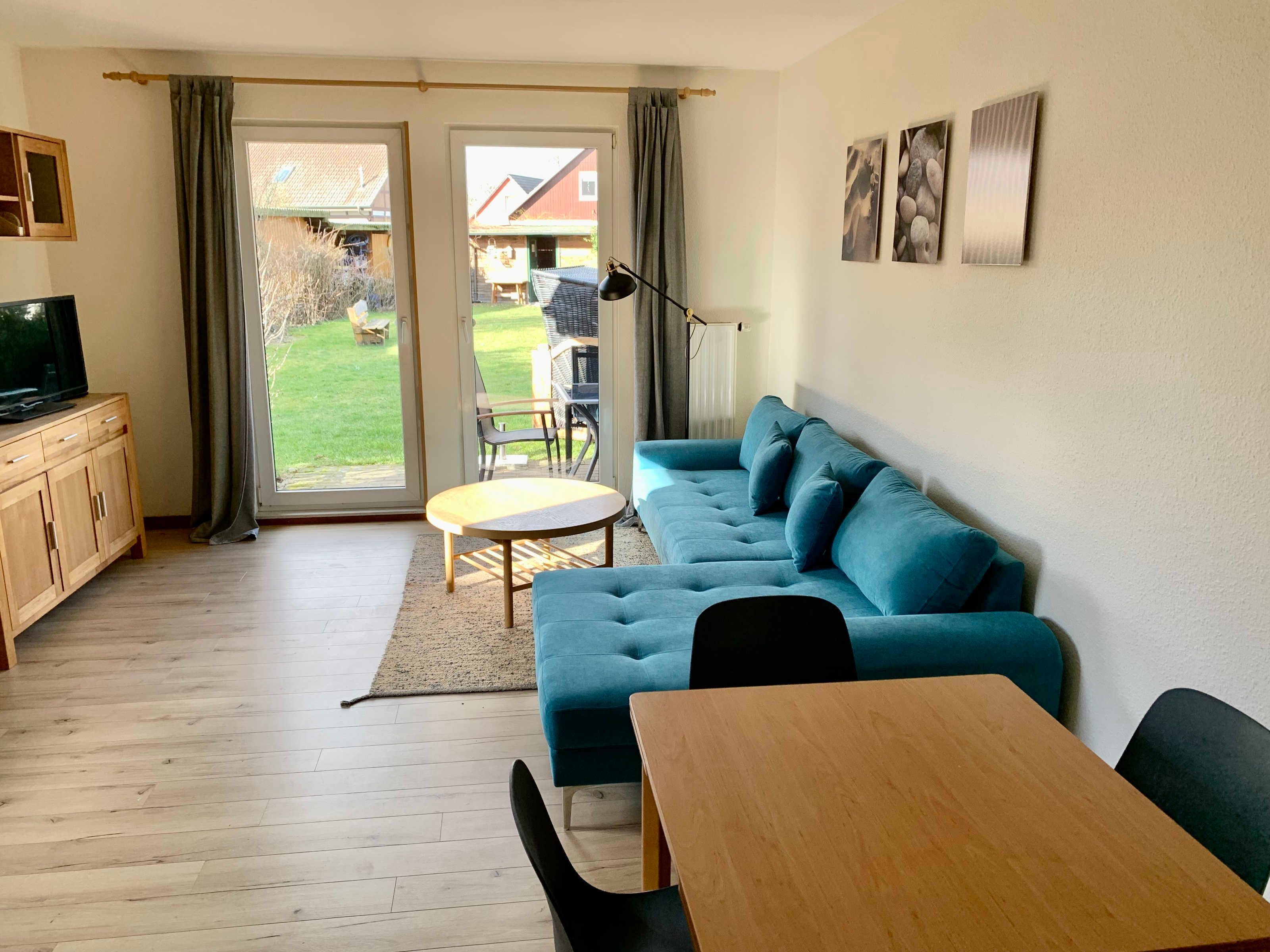Un salon avec un canapé bleu, une table en bois, un téléviseur posé sur un meuble et une vue sur un jardin, visible à travers une porte coulissante.