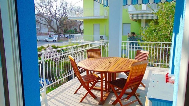 Un balcon avec une table et des chaises en bois rondes, une balustrade blanche et une vue sur un immeuble verdoyant.