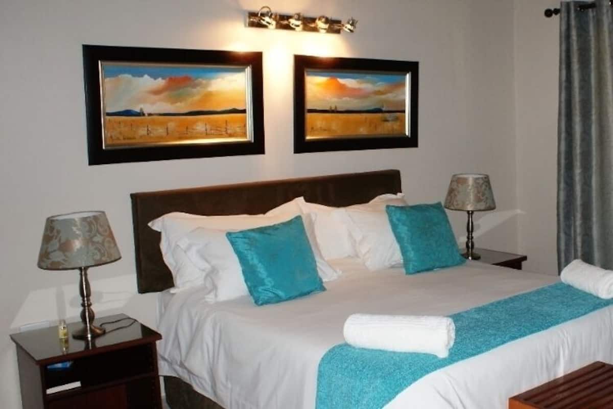 Una cama bien hecha con ropa de cama blanca y azul, dos lámparas de noche y dos cuadros de paisajes en la pared.