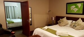 A hotel room with a bed, a chair, a desk, a lamp, and a painting on the wall. - Gabbata Lodge (Roodeplaat)